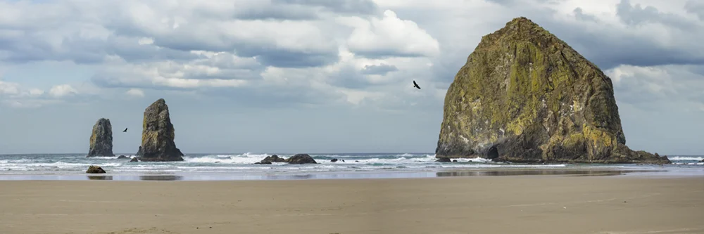 A panoramic landscape view of Haystack rock as viewed from the beach on a cloudy day